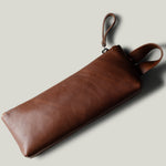A brown leather zip pouch with a wrist strap, shown at an angle on a light gray background with a soft shadow.