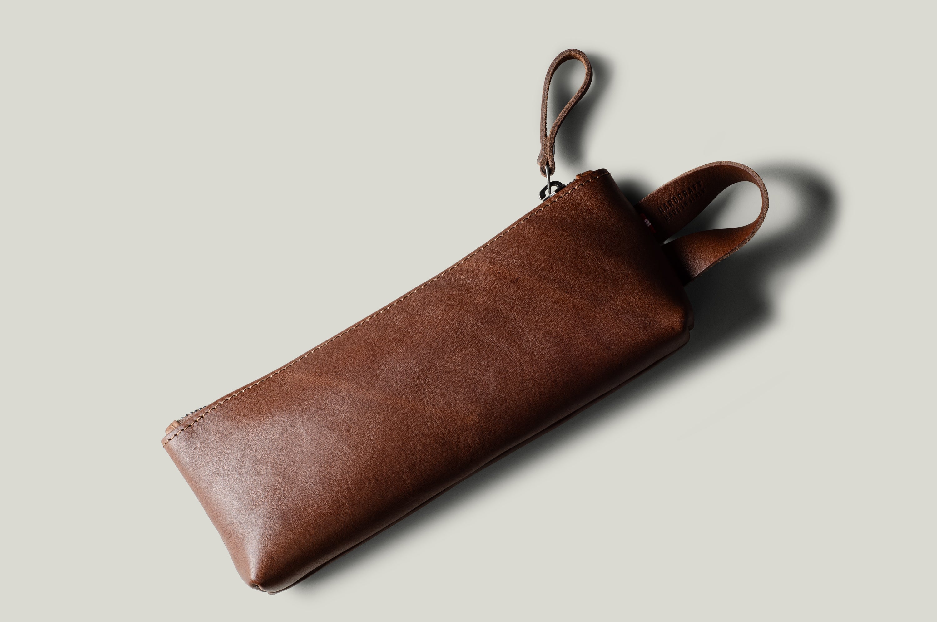 A brown leather zip pouch with a wrist strap, shown at an angle on a light gray background with a soft shadow.
