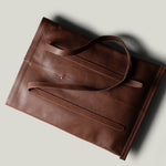 A brown leather tote bag with a minimalist design, photographed from an overhead angle on a light gray background with a soft shadow.