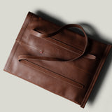 A brown leather tote bag with a minimalist design, photographed from an overhead angle on a light gray background with a soft shadow.
