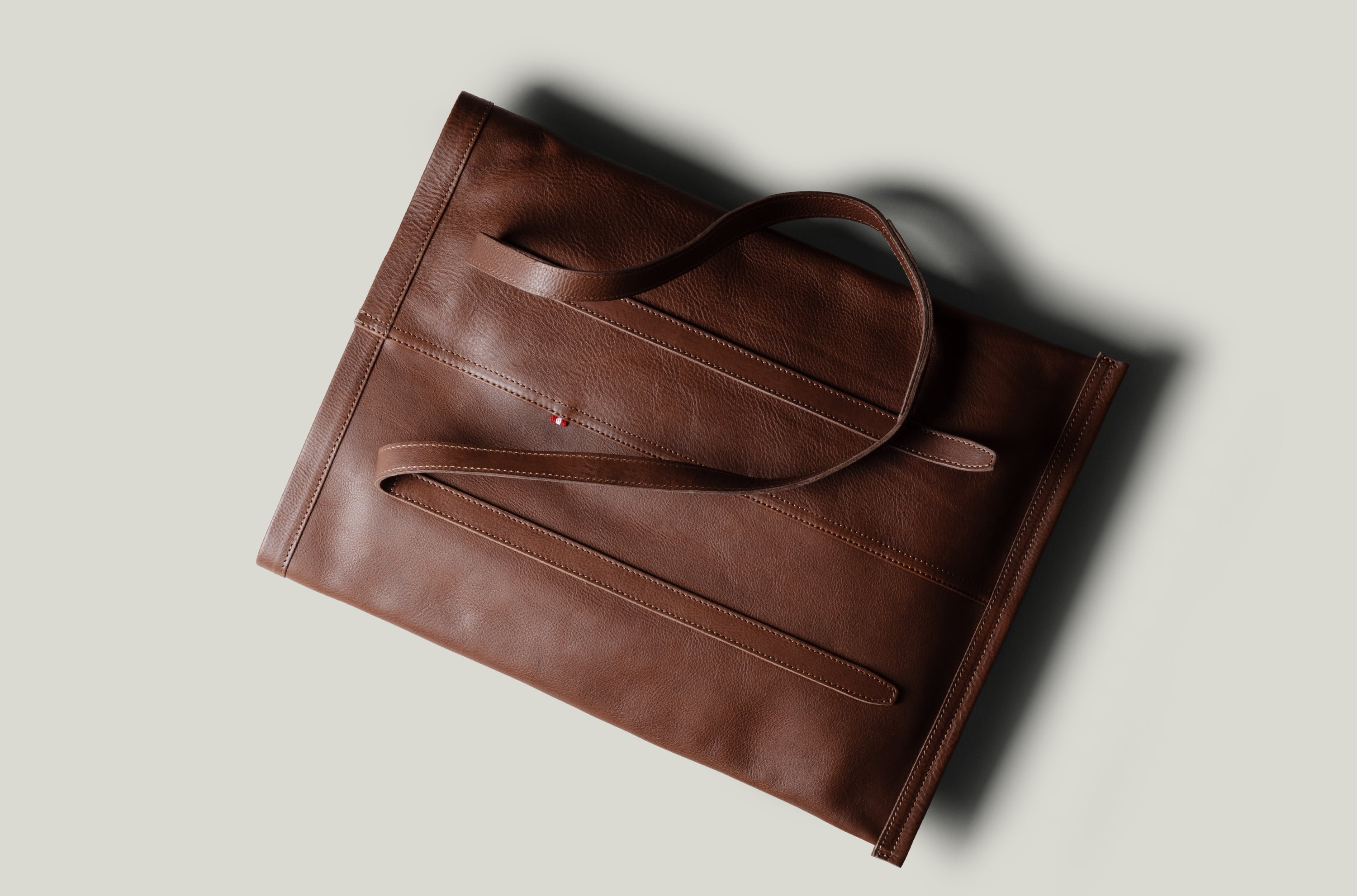 A brown leather tote bag with a minimalist design, photographed from an overhead angle on a light gray background with a soft shadow.