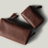 Two brown pebbled leather zip pouches, shown on a light gray background. One larger pouch is positioned diagonally in the foreground, and a smaller matching pouch is behind it.