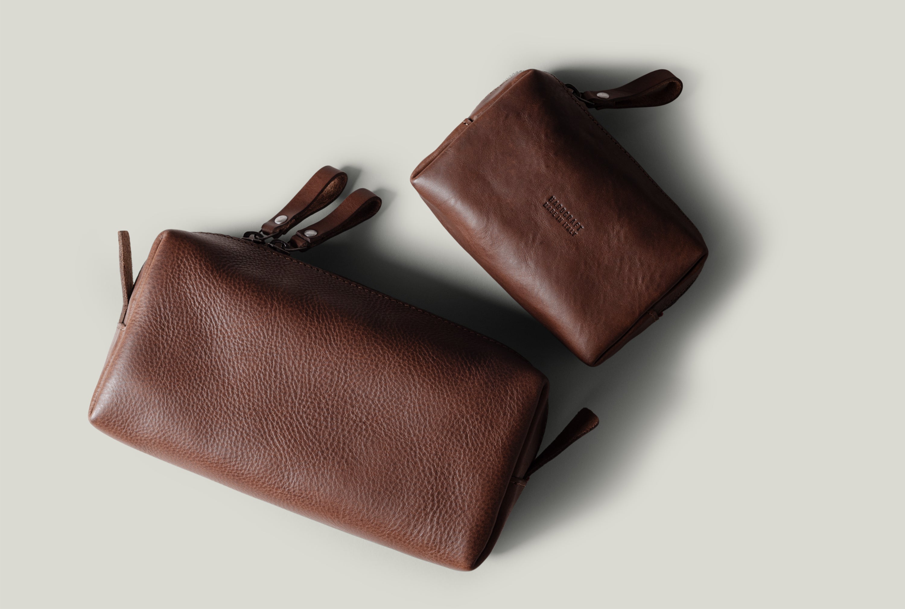 Two brown pebbled leather zip pouches, shown on a light gray background. One larger pouch is positioned diagonally in the foreground, and a smaller matching pouch is behind it.