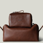Two brown pebbled leather toiletry bags stacked together on a light gray background. The smaller rectangular zip pouch sits on top of a larger pouch.