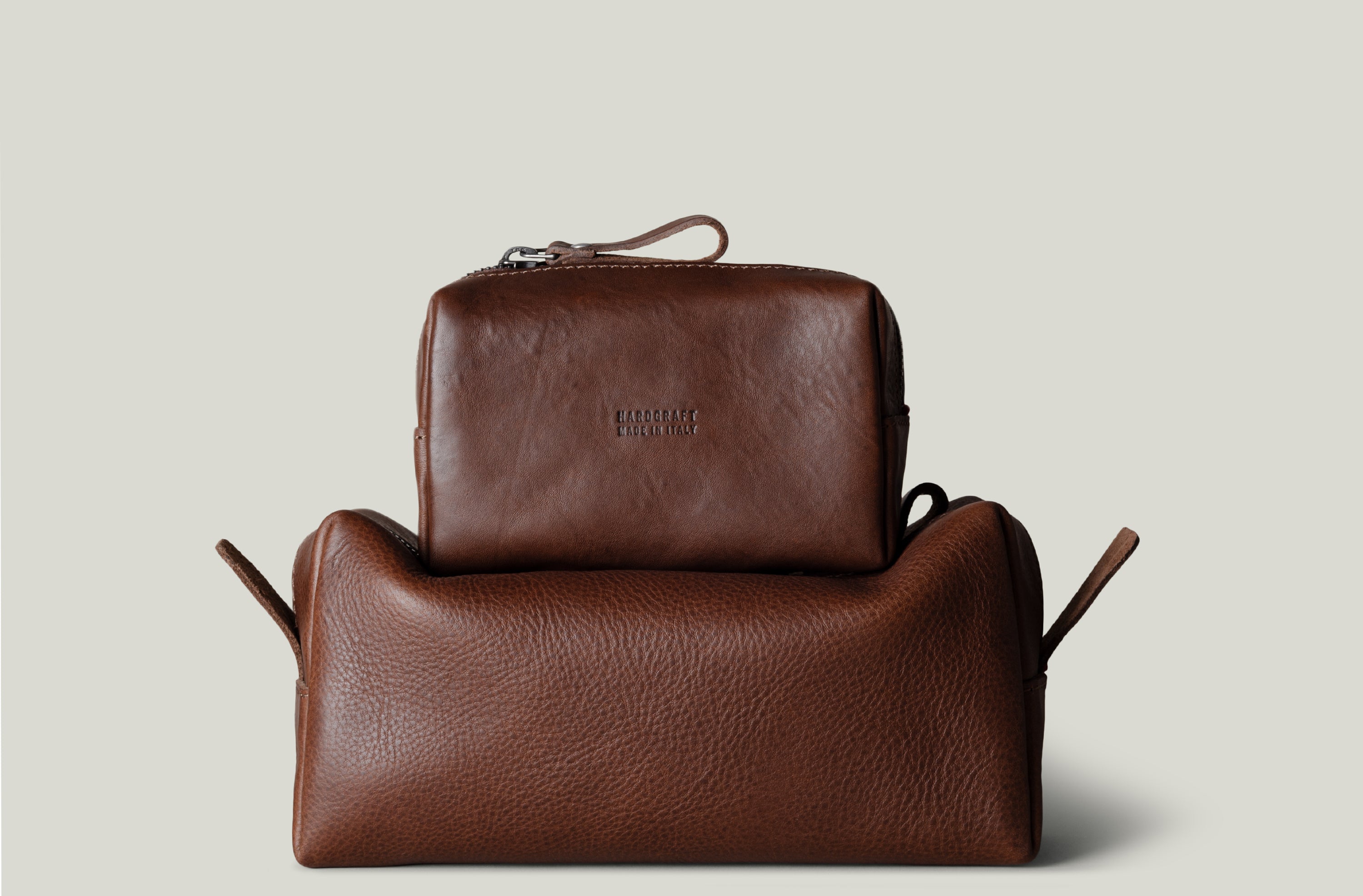 Two brown pebbled leather toiletry bags stacked together on a light gray background. The smaller rectangular zip pouch sits on top of a larger pouch.