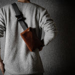 Person wearing a gray sweater with a brown leather bag slung over one shoulder against a dark background