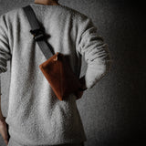 Person wearing a gray sweater with a brown leather bag slung over one shoulder against a dark background