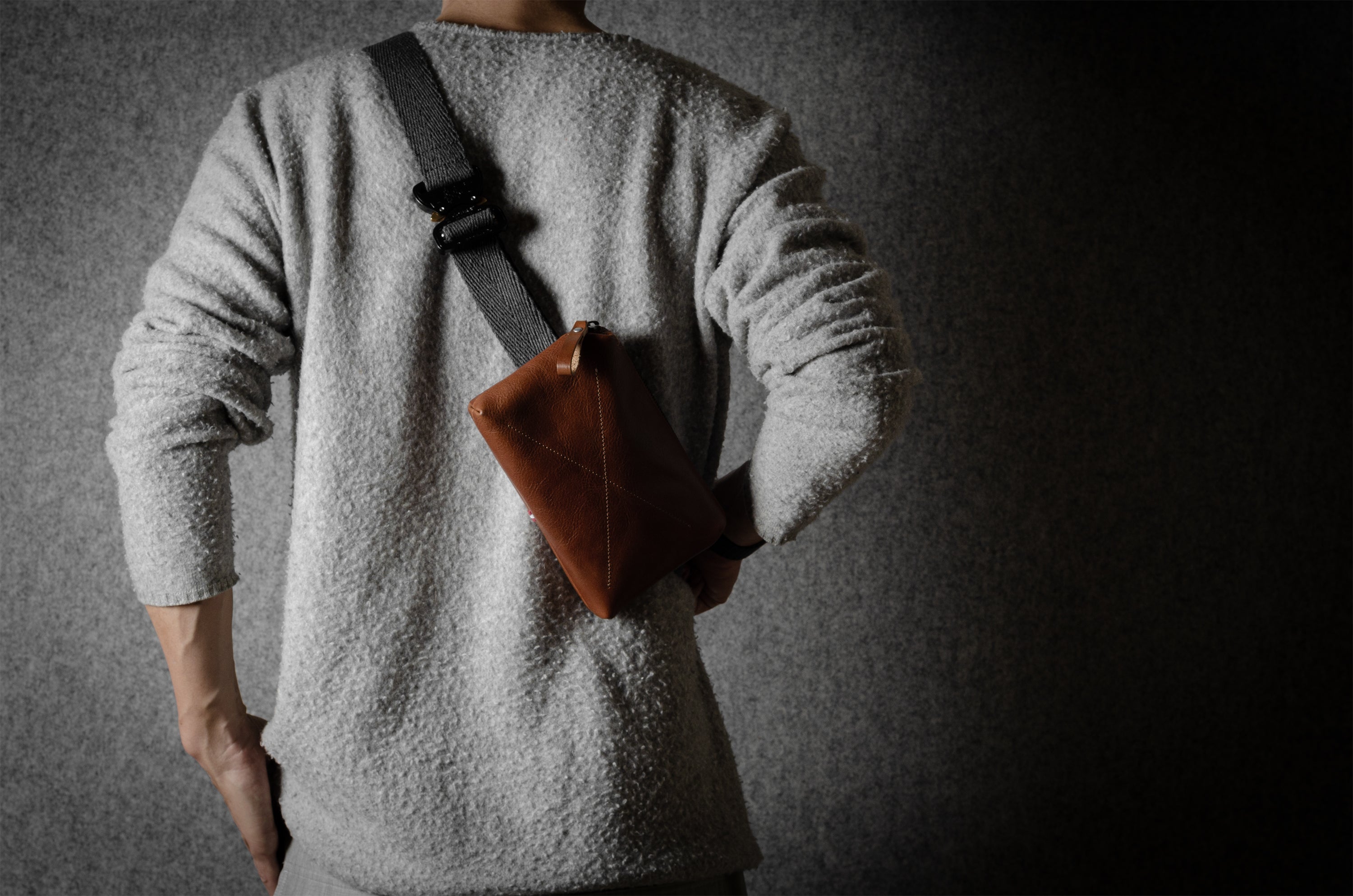 Person wearing a gray sweater with a brown leather bag slung over one shoulder against a dark background