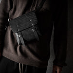 Person wearing a dark brown sweater with a black leather bag over one shoulder against a gray background