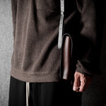 Person wearing a brown textured sweater holding a brown leather bag against a dark background