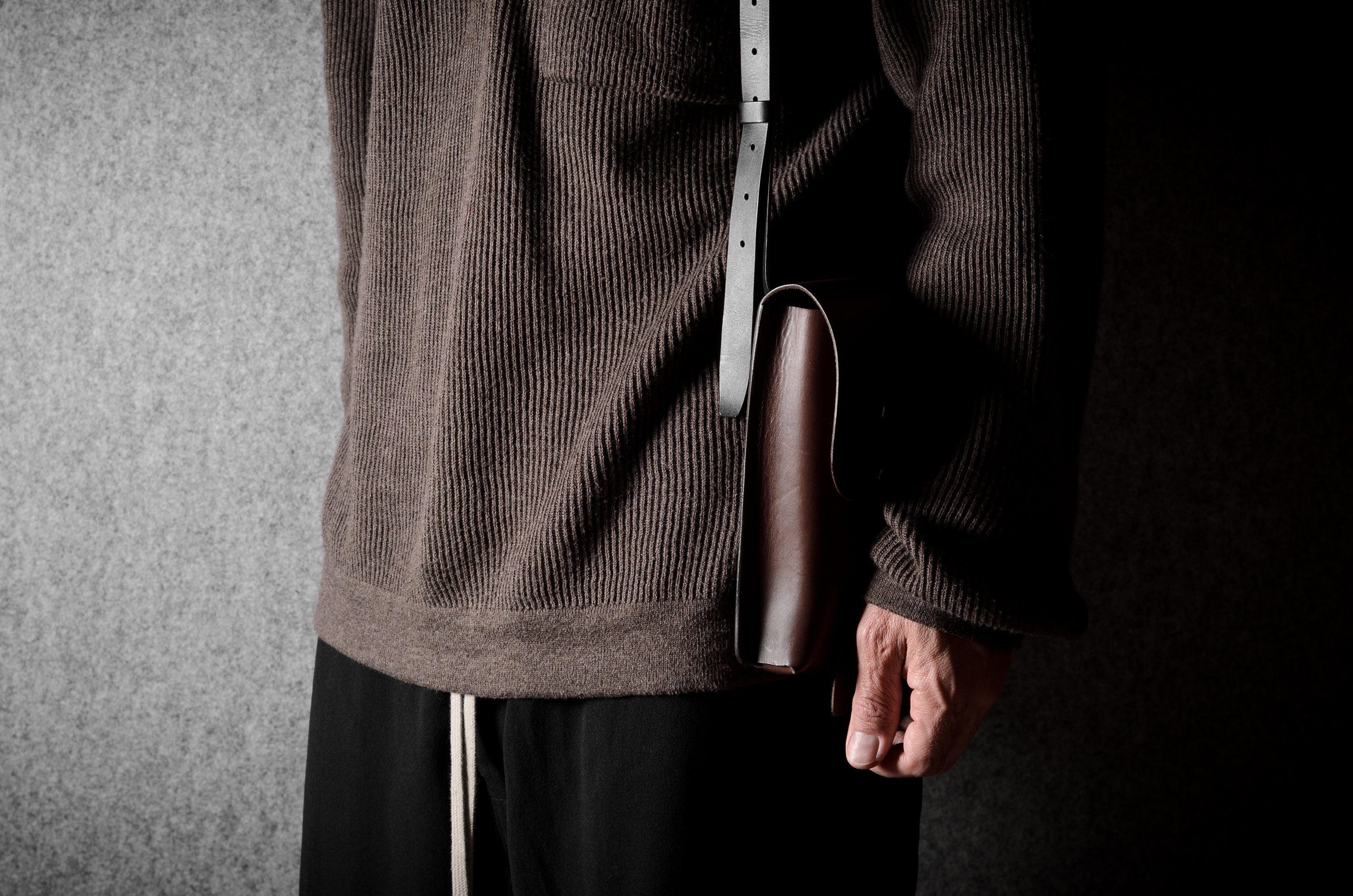 Person wearing a brown textured sweater holding a brown leather bag against a dark background