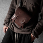 Person wearing a brown sweater with a brown leather satchel over one shoulder against a gray background