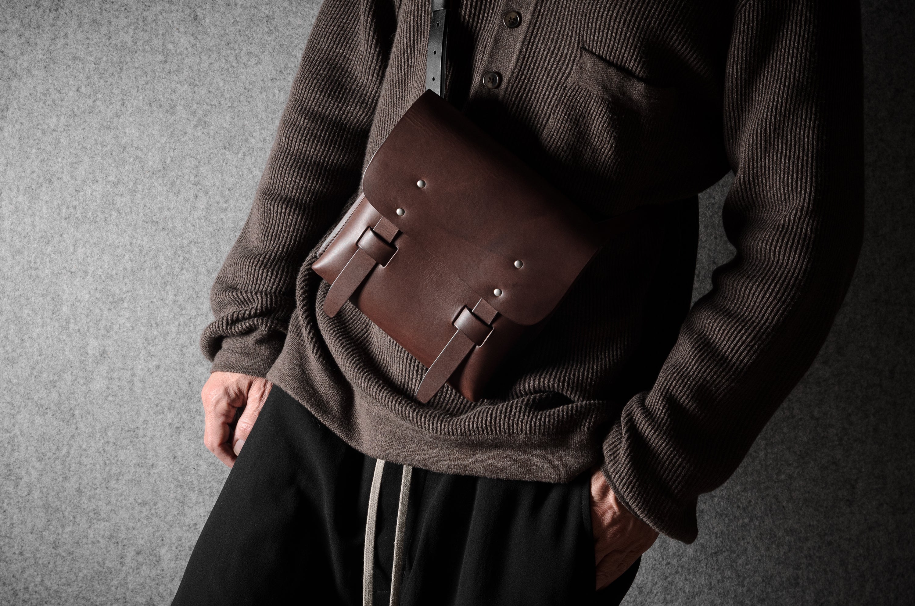 Person wearing a brown sweater with a brown leather satchel over one shoulder against a gray background