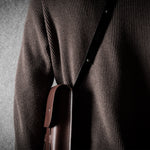 Person wearing a brown sweater with a leather bag over their shoulder against a gray background