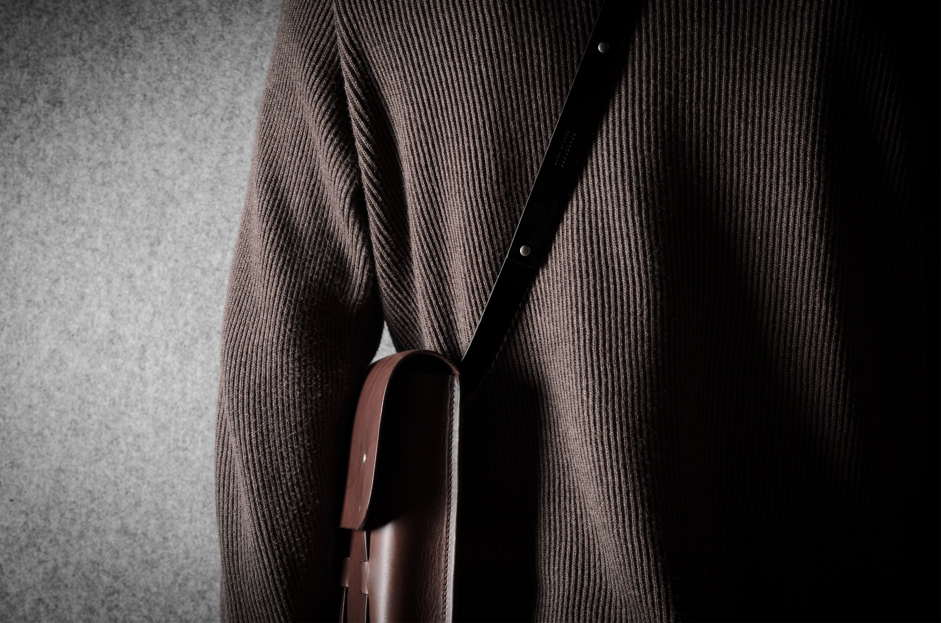 Person wearing a brown sweater with a leather bag over their shoulder against a gray background