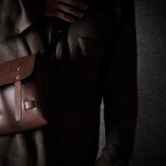 Brown leather satchel bag held by a person wearing a dark sweater against a dark background