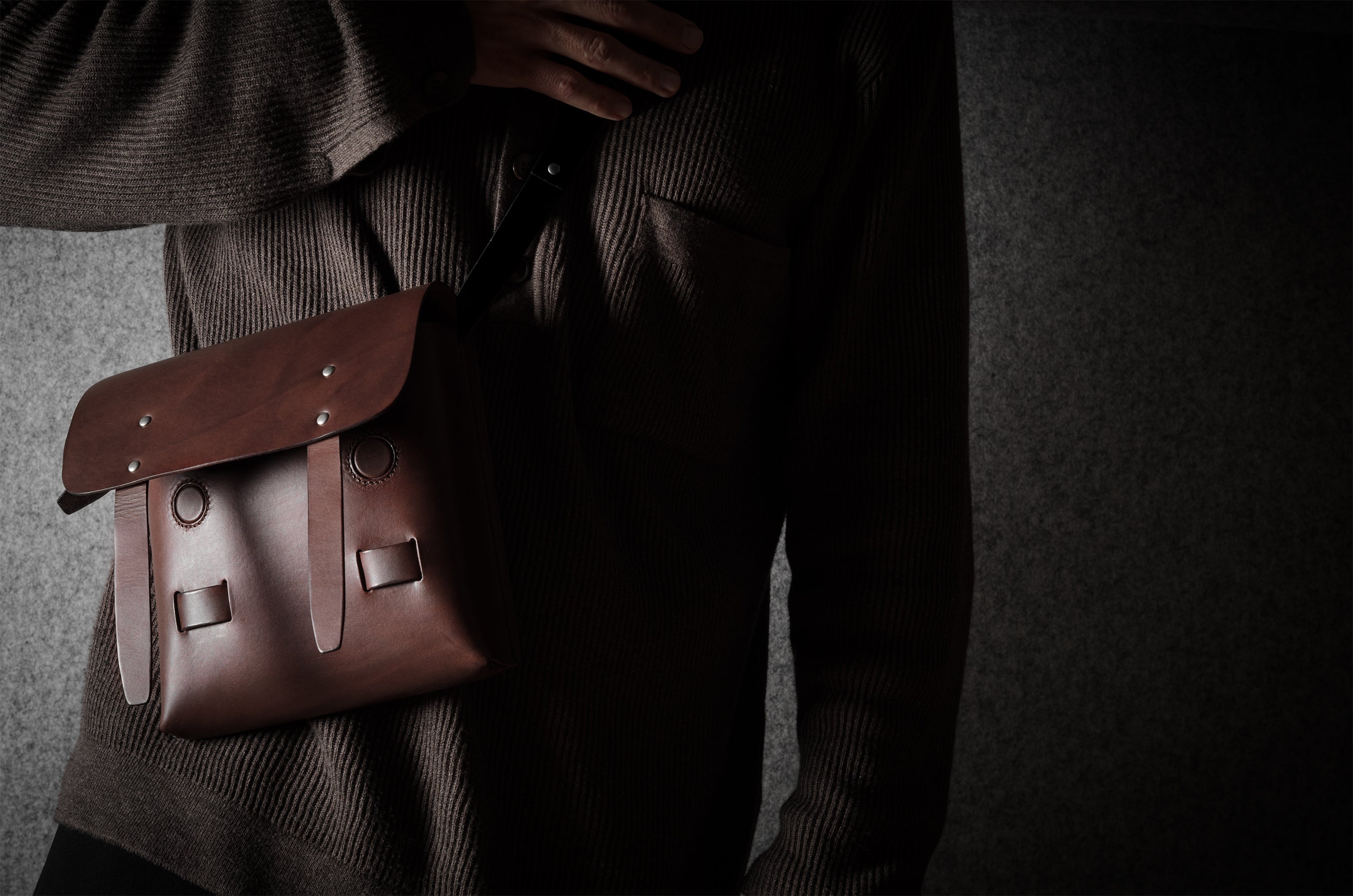 Brown leather satchel bag held by a person wearing a dark sweater against a dark background
