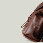 A brown leather backpack with a flap top, front pocket, and adjustable shoulder strap, photographed on a plain light gray background.