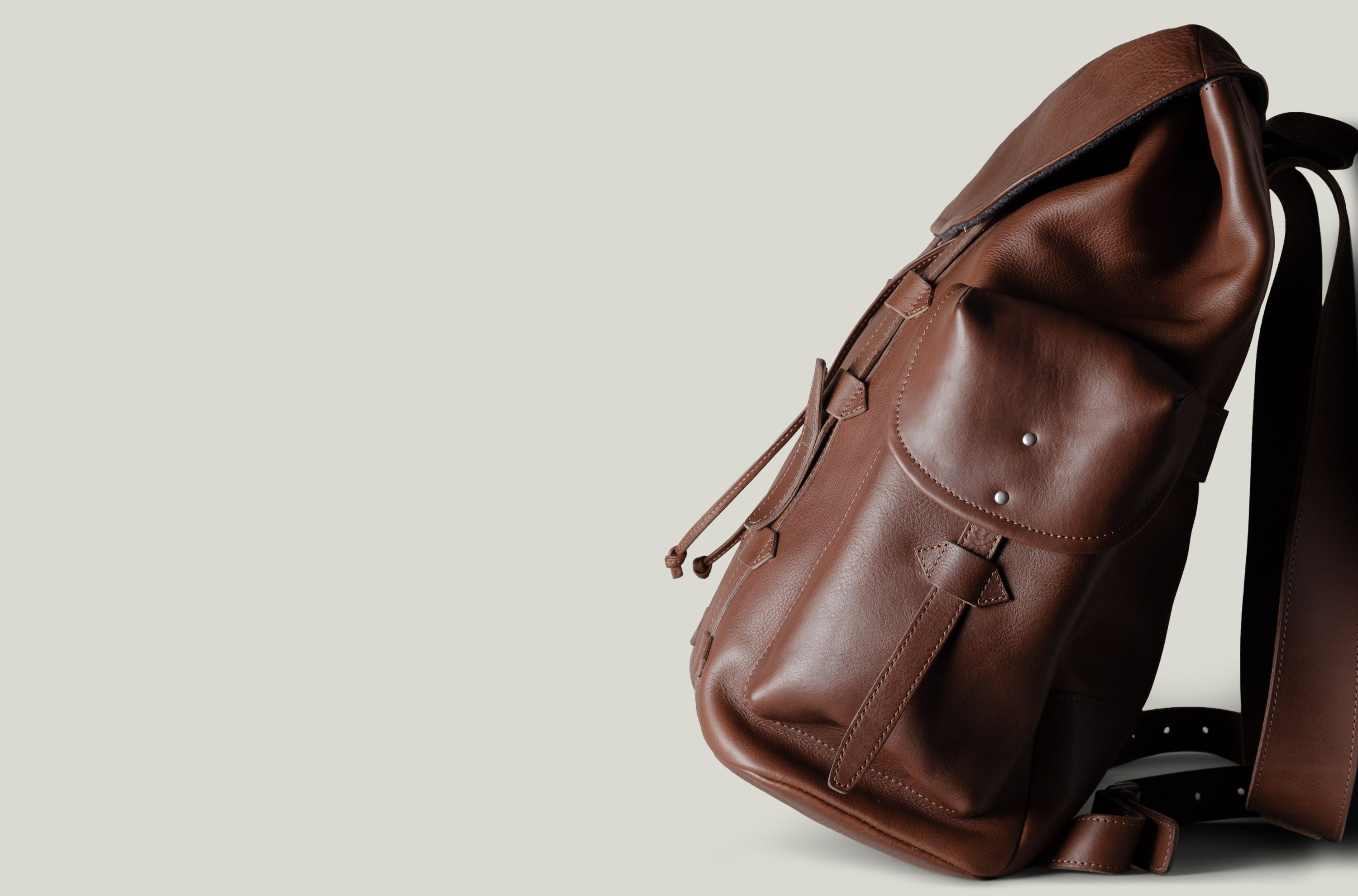 A brown leather backpack with a flap top, front pocket, and adjustable shoulder strap, photographed on a plain light gray background.