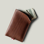 A brown textured leather card holder wallet with a small red tab, shown open with folded dollar bills partially sticking out, on a light gray background.