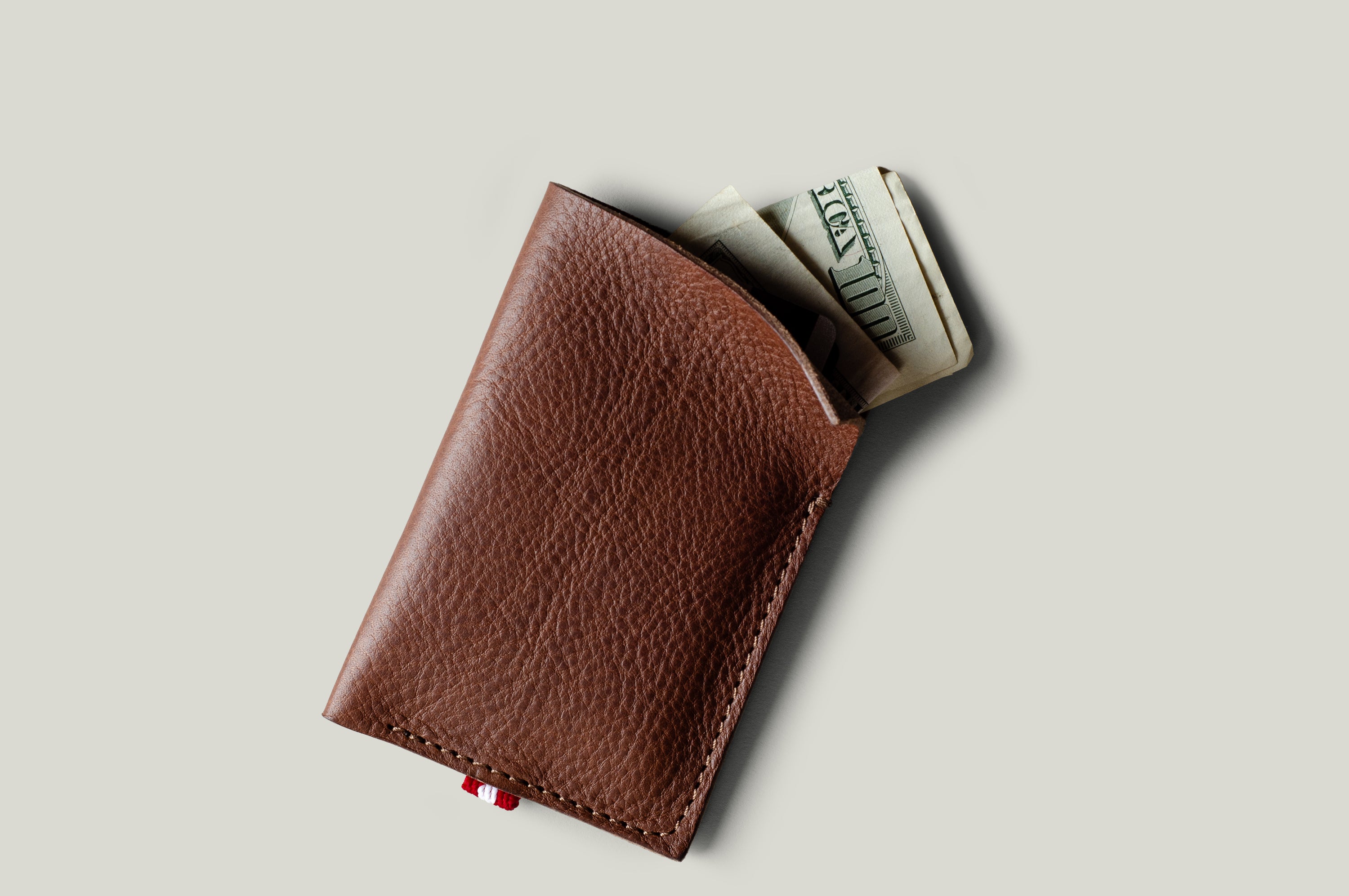 A brown textured leather card holder wallet with a small red tab, shown open with folded dollar bills partially sticking out, on a light gray background.