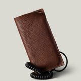 A brown textured leather iPhone 18 Pro case with a black coiled cord, shown standing upright on a light gray background.