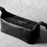 A black textured leather toiletry pouch with a top zipper and adjustable strap, photographed on a gray felt surface.