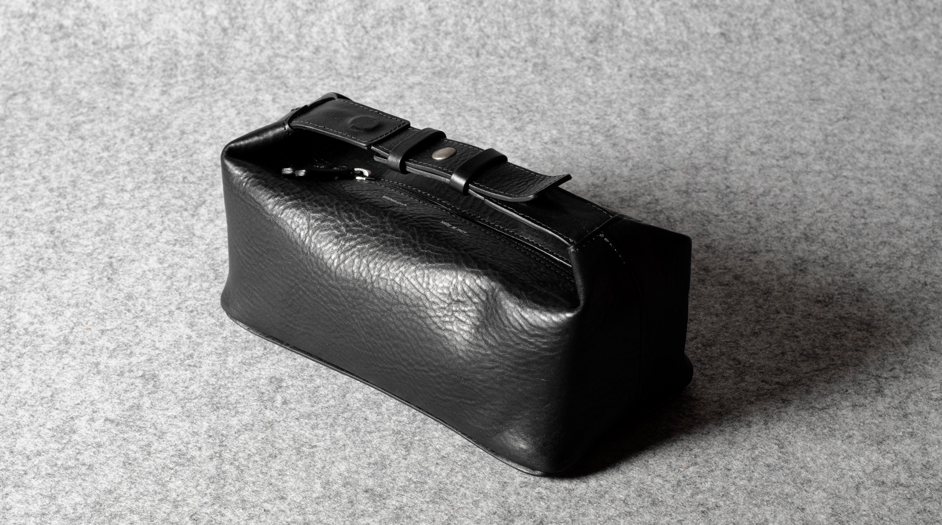 A black textured leather toiletry pouch with a buckle strap closure, photographed on a gray felt surface.