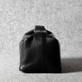 A black leather toiletry bag with a structured, rounded rectangular shape and a top handle, photographed from the back against a textured gray background.