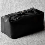 A black leather toiletry case with a strap and buckle closure, photographed on a textured gray surface.