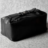 A black leather toiletry case with a strap and buckle closure, photographed on a textured gray surface.