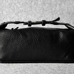 A black textured leather toiletry case with a top zipper and a thin strap detail, photographed on a neutral gray textured background.