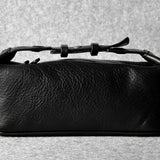A black textured leather toiletry case with a top zipper and a thin strap detail, photographed on a neutral gray textured background.