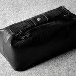 A black leather zippered pouch or toiletry case with a flap closure and metal snap detail, photographed on a textured gray surface.