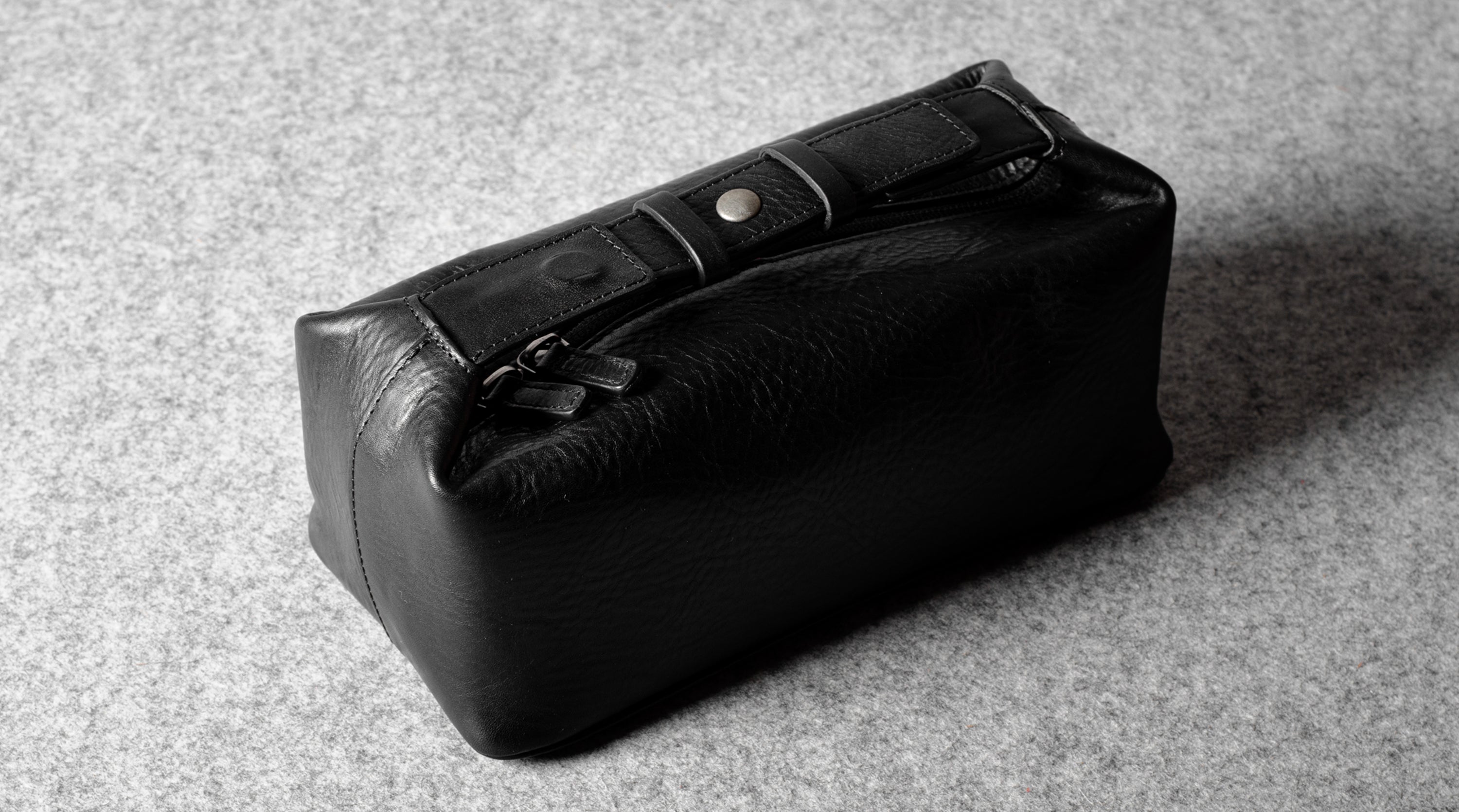 A black leather zippered pouch or toiletry case with a flap closure and metal snap detail, photographed on a textured gray surface.