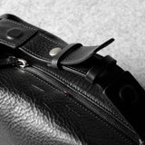 Close-up of a black textured leather toiletry bag with a zipper, snap-button strap, and metal hardware, photographed on a light gray surface.