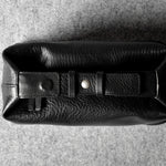 A black pebbled leather toiletry pouch with a buckle strap closure, photographed from a top-down angle on a textured gray background.