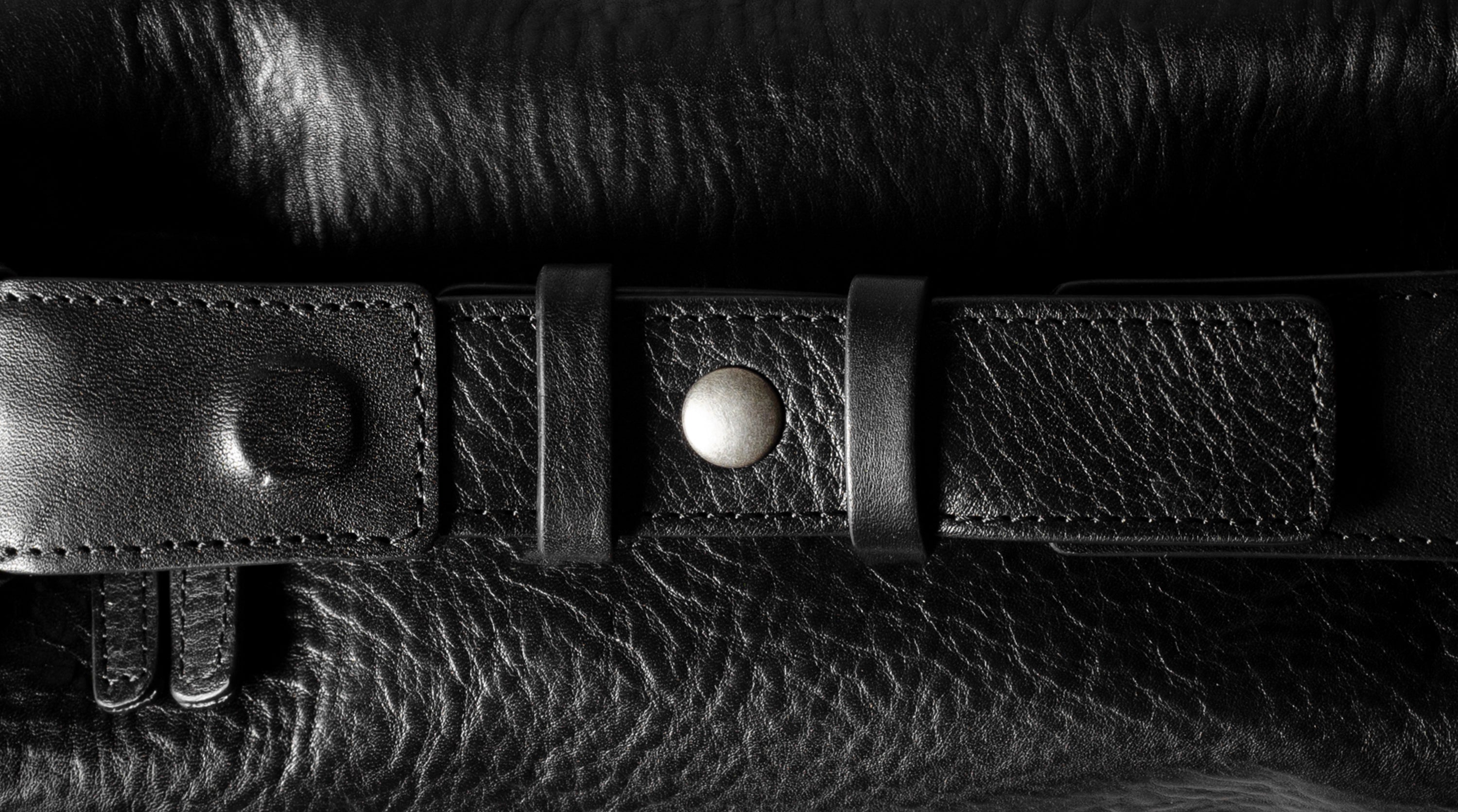 A close-up of a black leather belt with a silver-toned snap button and textured grain, laid horizontally across a matching black leather surface.