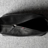 A black textured leather zippered toiletry pouch or small bag, shown from a top-down view on a gray felt surface.