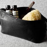 A black textured leather zippered toiletry pouch containing two small dark bottles, a brown wooden tool, and a yellow sponge, set on a gray felt surface.