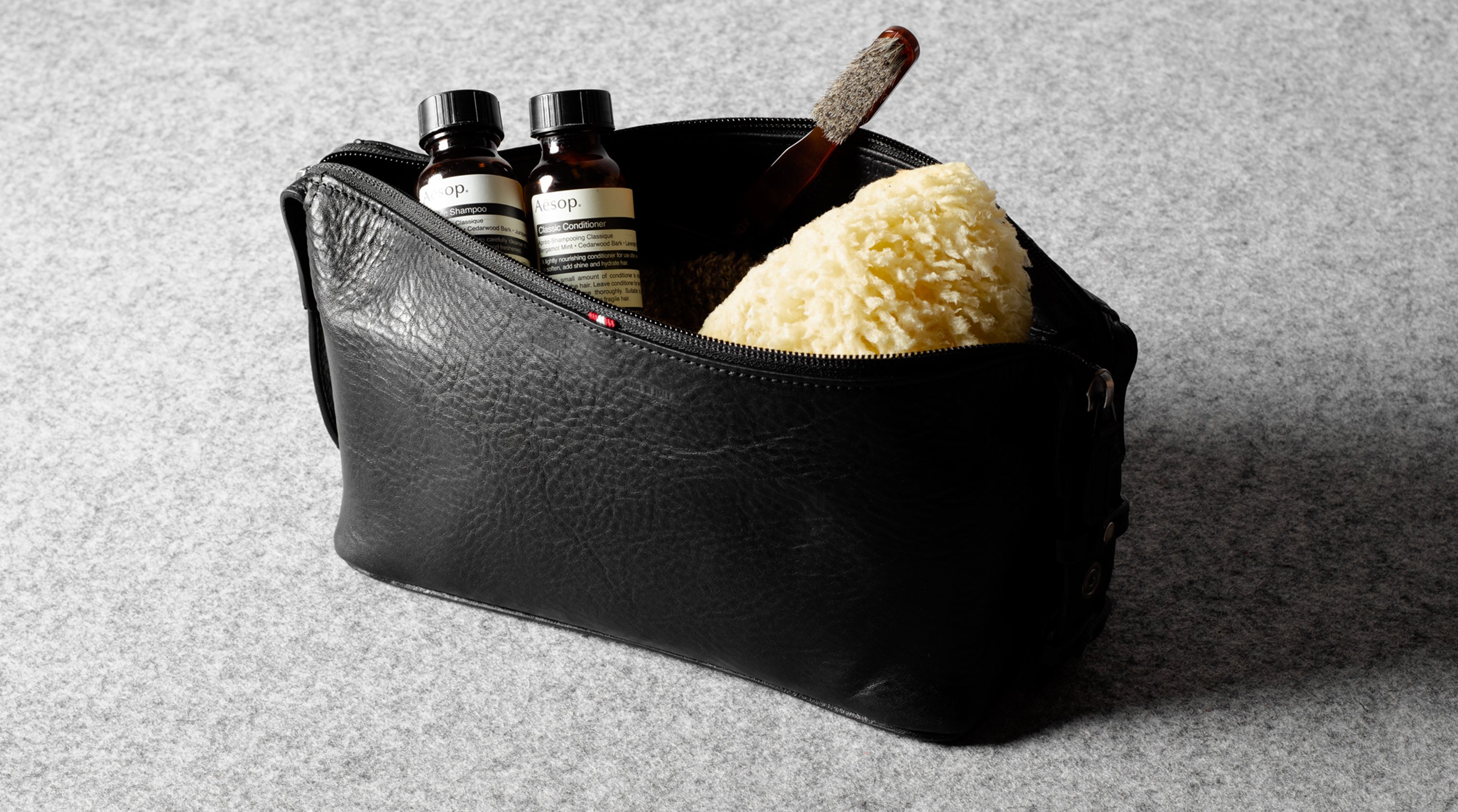 A black textured leather zippered toiletry pouch containing two small dark bottles, a brown wooden tool, and a yellow sponge, set on a gray felt surface.