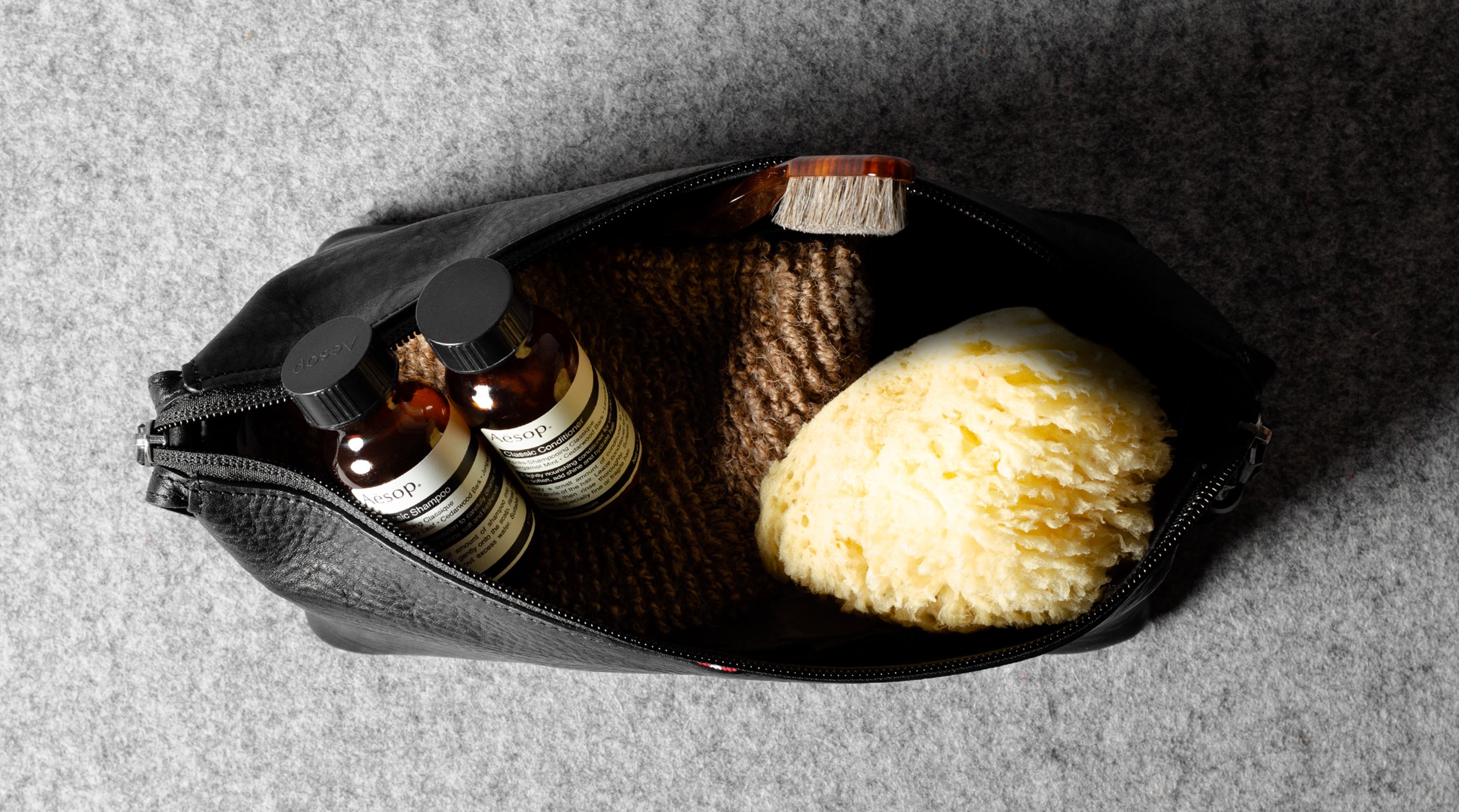 An open black zippered toiletry pouch on a gray textured background, containing two small amber bottles with black caps and white labels, a brown scrubber, and a yellow sponge.