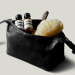 A black leather toiletry bag with a zipper and strap, shown open with two small striped bottles, a sponge, and a brush inside, on a light gray background.