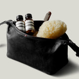A black leather toiletry bag with a zipper and strap, shown open with two small striped bottles, a sponge, and a brush inside, on a light gray background.