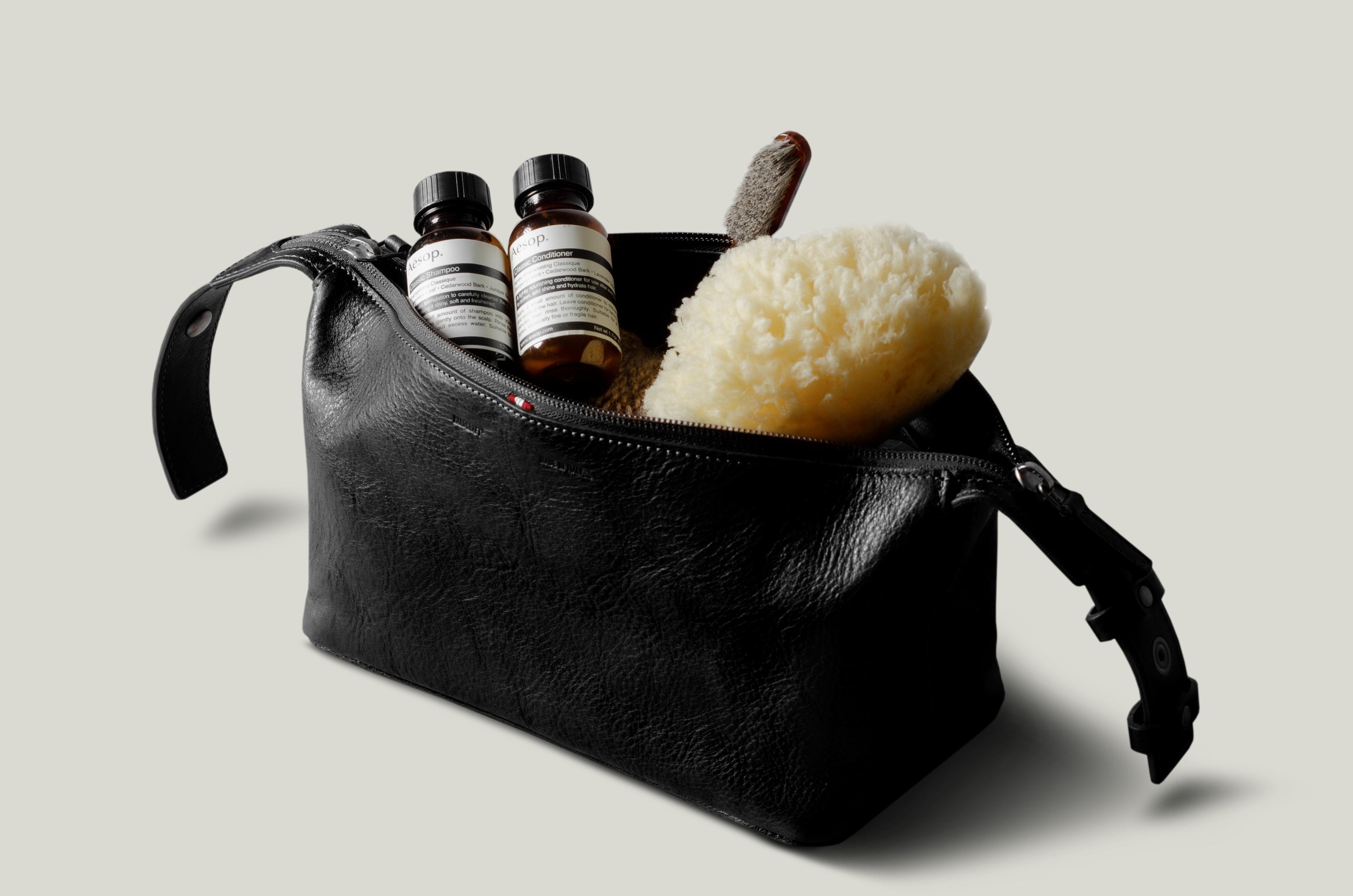 A black leather toiletry bag with a zipper and strap, shown open with two small striped bottles, a sponge, and a brush inside, on a light gray background.