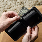 Person holding a black bi-fold wallet  with money on a wooden surface
