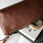 A brown leather zip pouch with a wrist strap, shown partially open and filled with stationery items like a notebook, tape, and pen, on a light textured background.