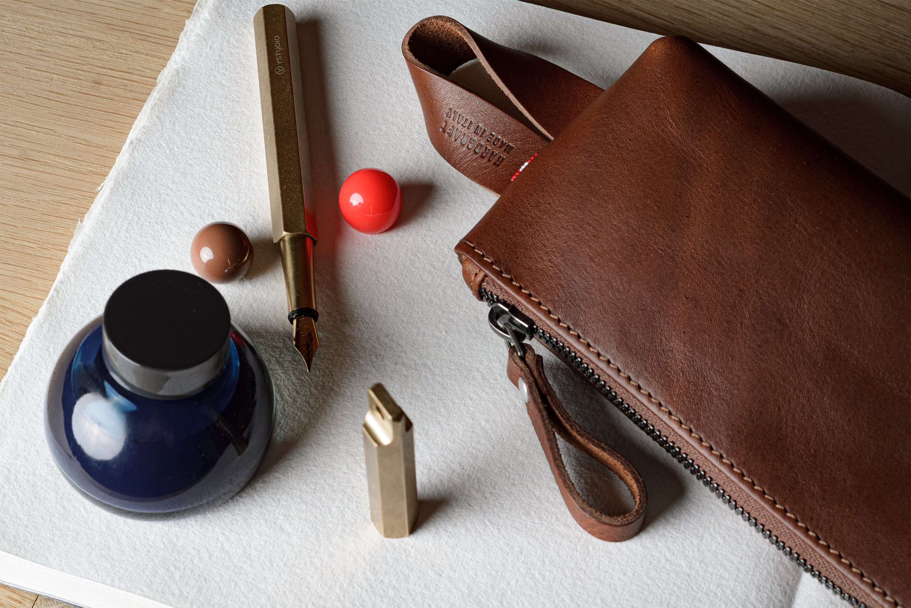 A brown leather zip pouch with a wrist strap, arranged on a white cloth alongside a gold pen, a blue ink bottle, a small red ball, and a gold lighter on a light wood surface.