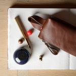 A brown leather zip pouch with a wrist strap, styled on a white sheet of paper over a light wood background alongside a gold pen, a small blue ink bottle, and decorative beads.