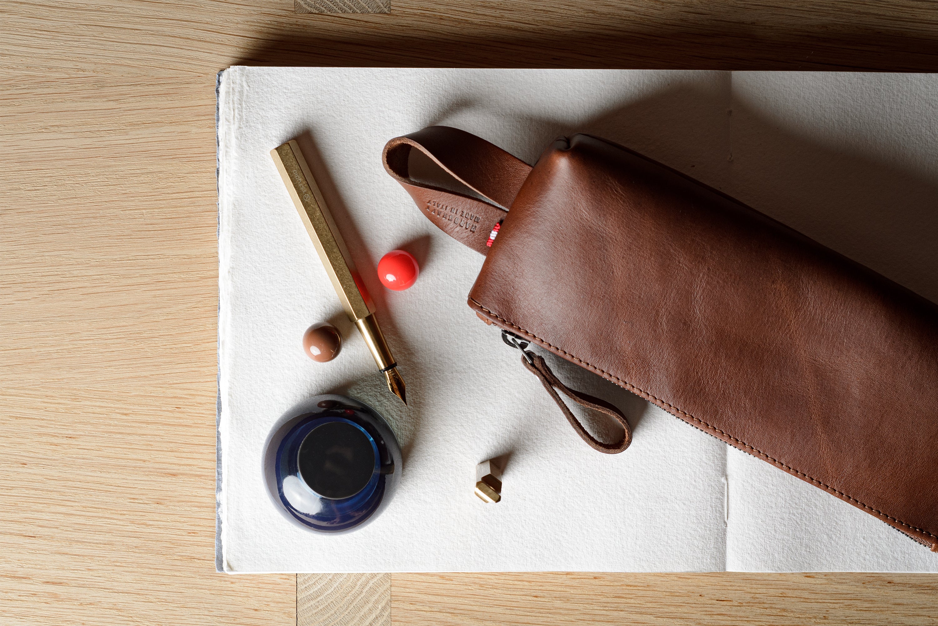 A brown leather zip pouch with a wrist strap, styled on a white sheet of paper over a light wood background alongside a gold pen, a small blue ink bottle, and decorative beads.